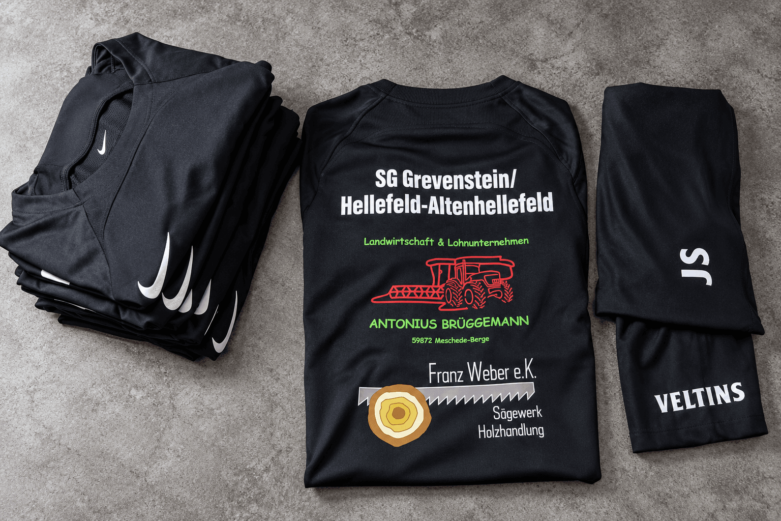 Gestapelte schwarze Vereinsoberteile mit großem Sponsoren-Rückenmotiven und Bedruckung der Hosen